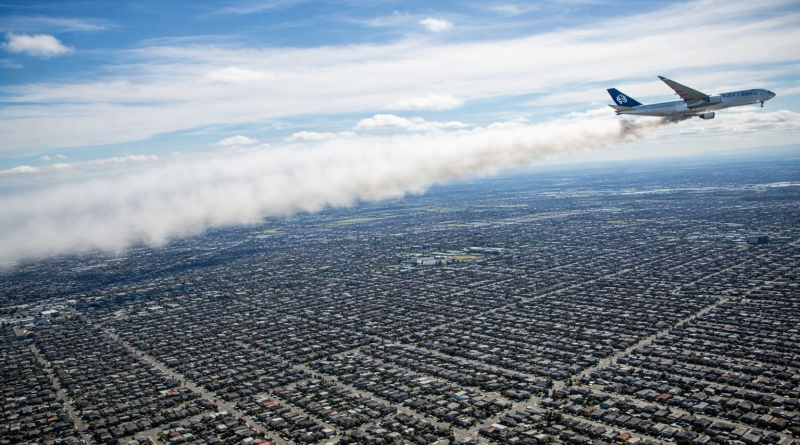 Avion en vol larguant un nuage de carburant au-dessus d’une vaste zone urbaine dense, avec des milliers d’habitations visibles en contrebas.