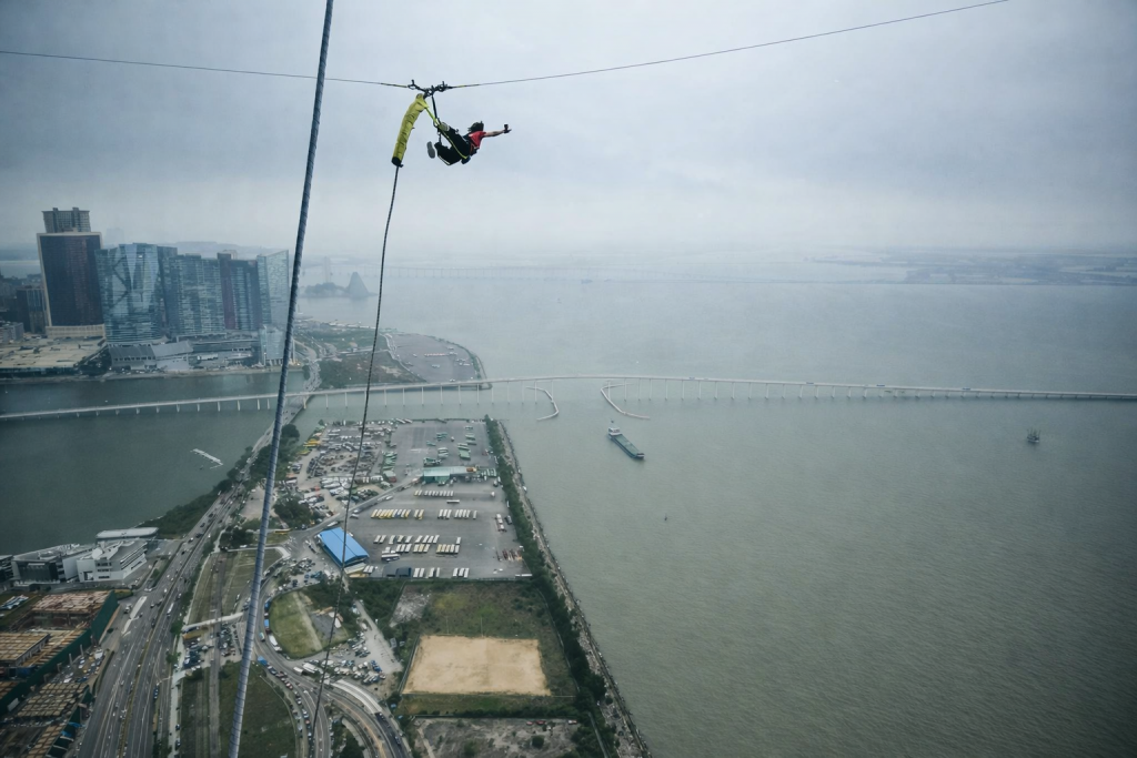 Personne suspendue à un câble en très grande hauteur au-dessus de l’eau, avec une ville et un pont visibles en contrebas, scène de saut extrême et vertigineuse.