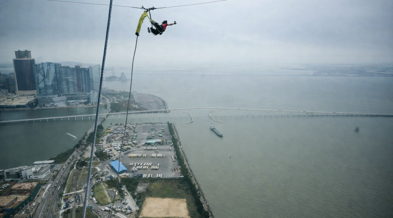 Personne suspendue à un câble en très grande hauteur au-dessus de l’eau, avec une ville et un pont visibles en contrebas, scène de saut extrême et vertigineuse.