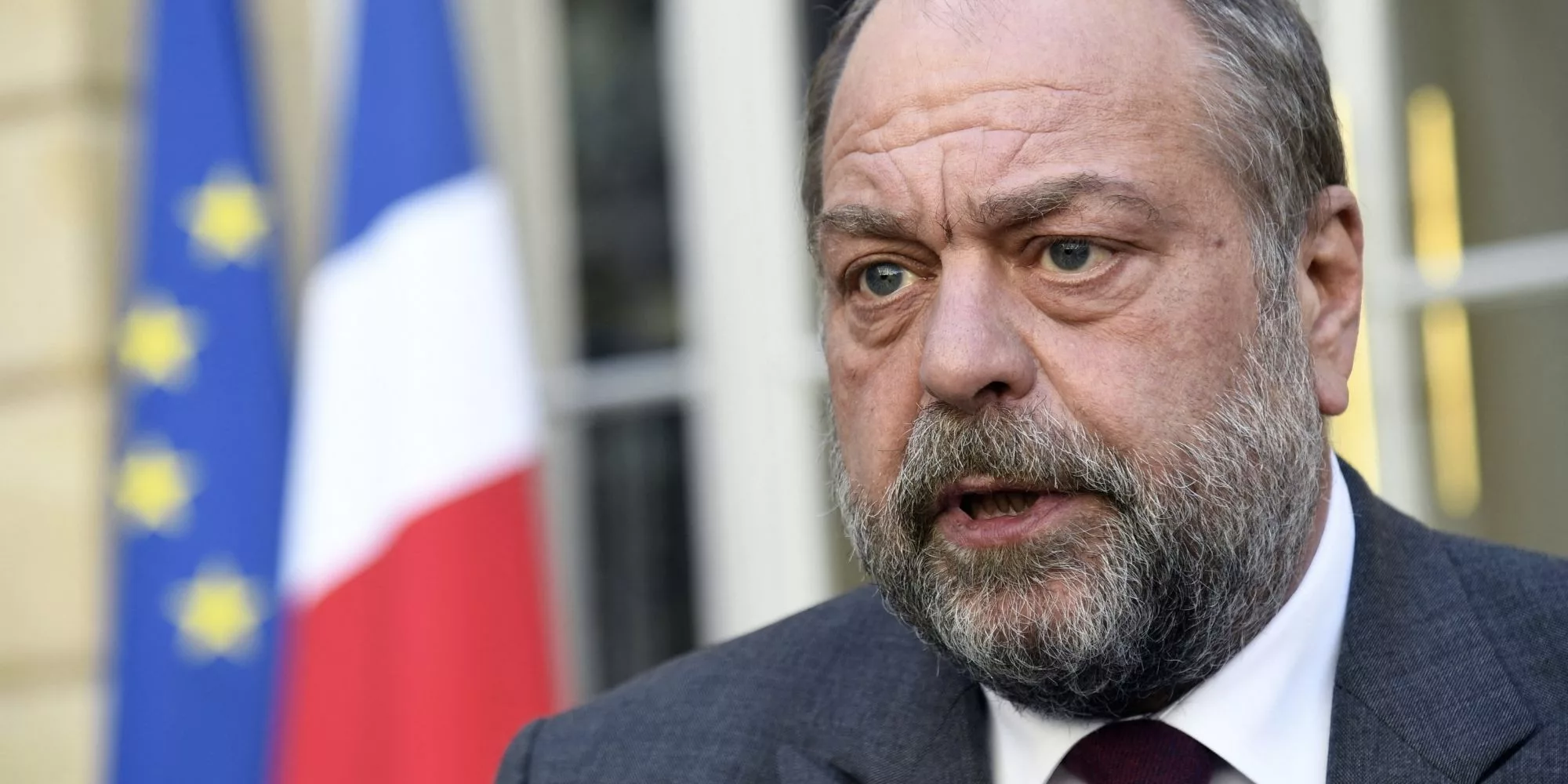 Portrait rapproché d’un homme en costume, au visage sérieux et marqué, parlant devant des drapeaux français et européens en arrière-plan.