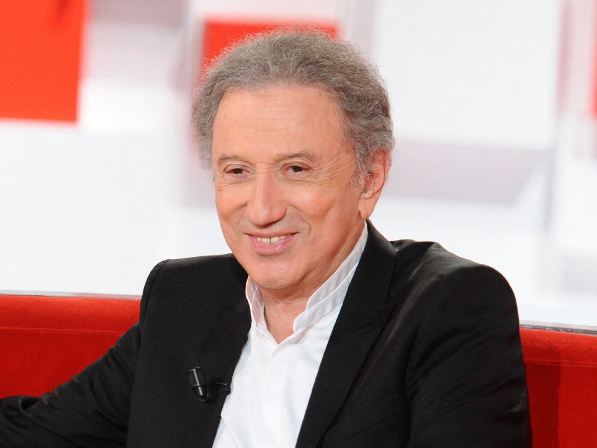 Michel Drucker assis sur un canapé rouge lors d’une émission télévisée, souriant, vêtu d’une chemise blanche et d’une veste noire, en studio lumineux.