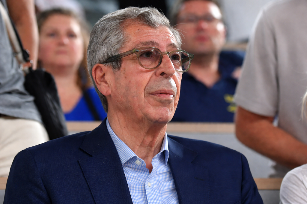 Homme âgé portant des lunettes, assis dans une salle, observant attentivement une scène devant lui, avec un public flou en arrière-plan.