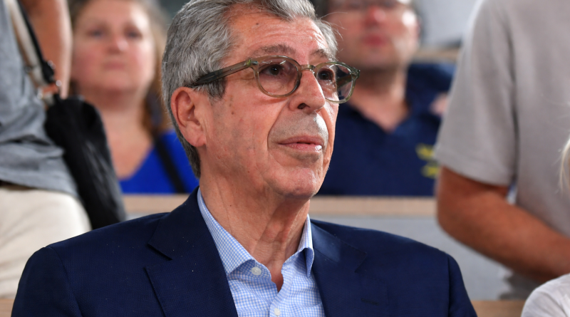 Homme âgé portant des lunettes, assis dans une salle, observant attentivement une scène devant lui, avec un public flou en arrière-plan.