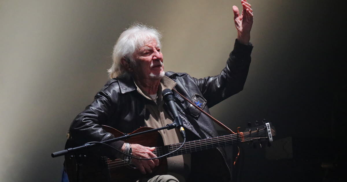 Hugues Aufray sur scène, guitare à la main, visage concentré, dans une ambiance de concert