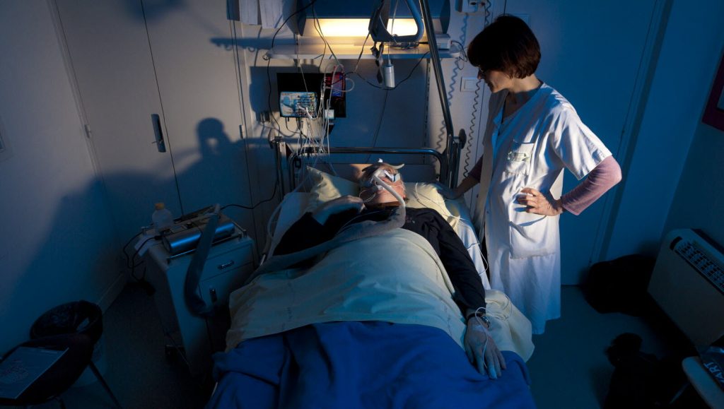 Patient allongé dans un lit d’hôpital, relié à de nombreux capteurs et porteur d’un masque respiratoire, tandis qu’une soignante en blouse blanche se tient à son chevet dans une chambre éclairée en bleu