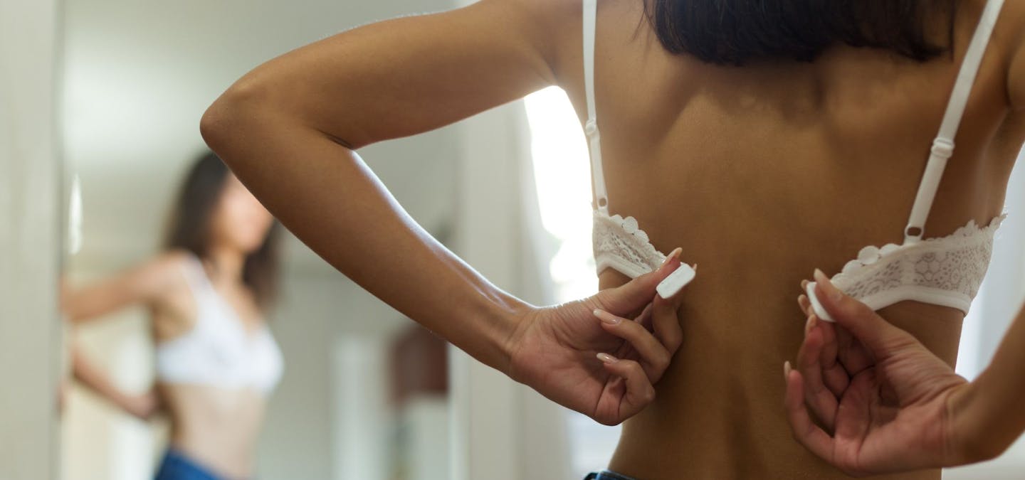 Femme en train d’attacher un soutien-gorge blanc devant un miroir, symbolisant la réflexion sur le port ou l’abandon du soutien-gorge et ses effets sur la poitrine.