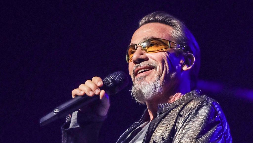 Chanteur sur scène, souriant et tenant un micro sous des projecteurs violets, vêtu d’une veste brillante et portant des lunettes teintées.