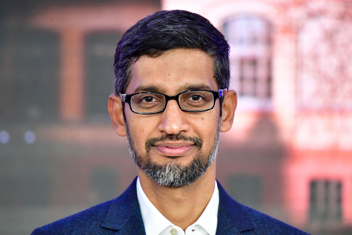 Portrait d’un homme portant des lunettes et une barbe soignée, posé face caméra, avec un léger sourire et un fond flou aux tons chauds.