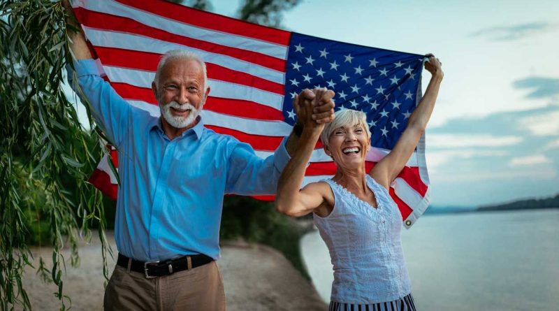 Un couple de retraités souriants marche main dans la main au bord d’un lac, tenant un grand drapeau américain derrière eux, symbole de leur expatriation heureuse en France.
