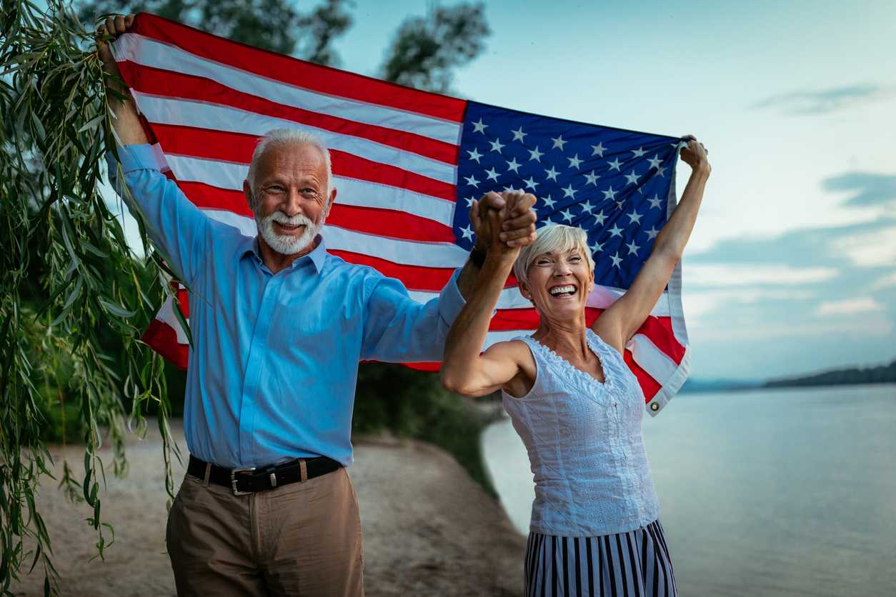 Un couple de retraités souriants marche main dans la main au bord d’un lac, tenant un grand drapeau américain derrière eux, symbole de leur expatriation heureuse en France.