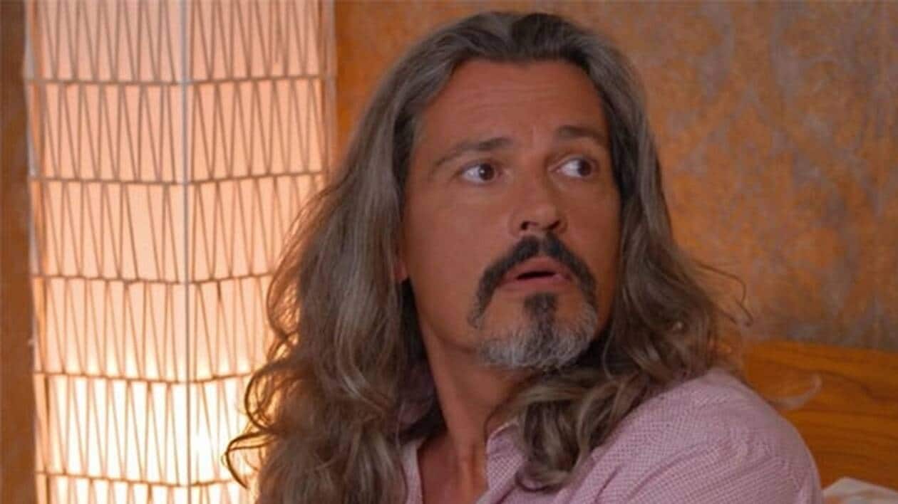 Homme aux cheveux longs et grisonnants assis dans une pièce éclairée par une lampe, le visage surpris tourné vers la droite.