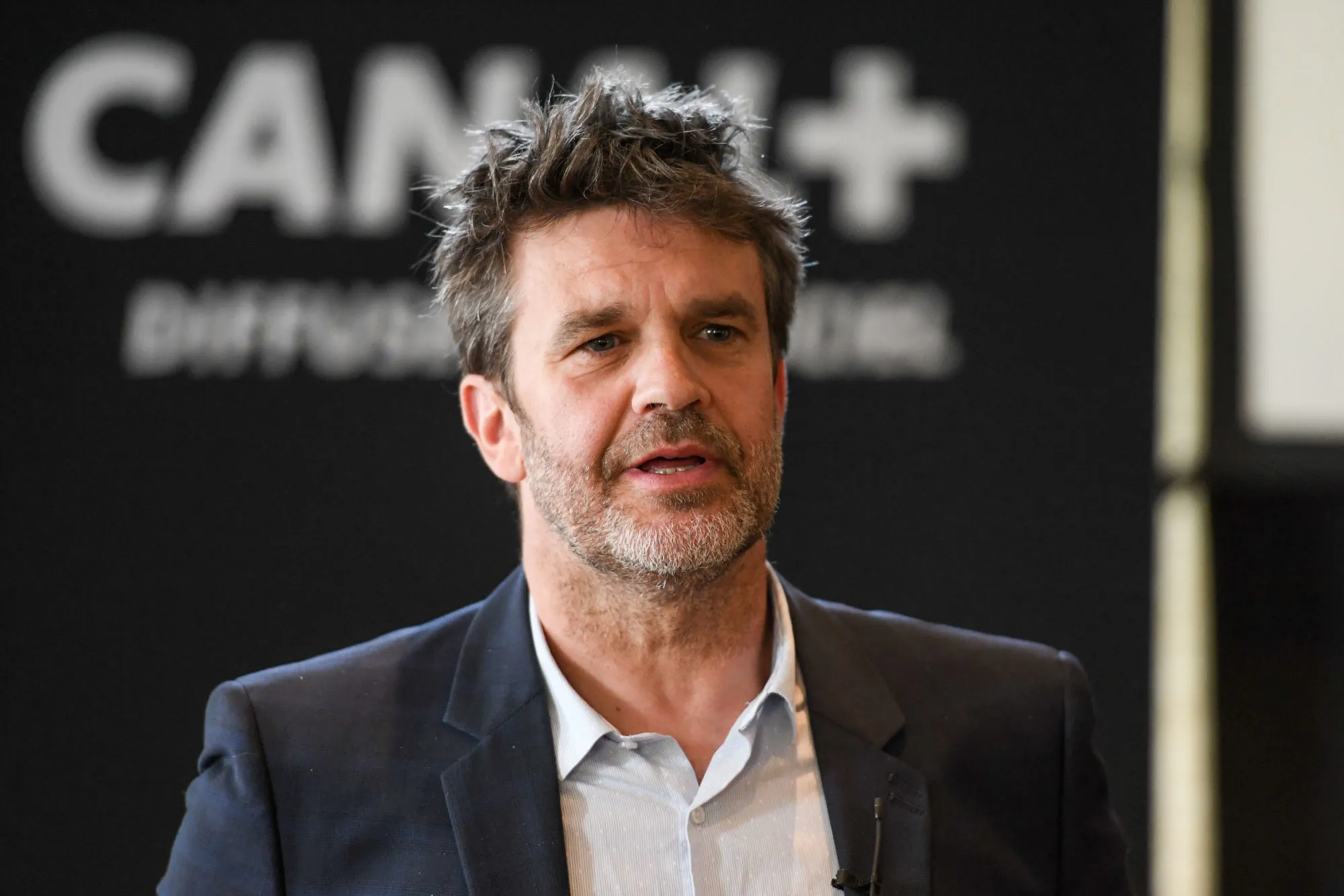 Homme en costume parlant lors d’un événement médiatique, debout devant un fond sombre où apparaît le logo d’une chaîne de télévision.