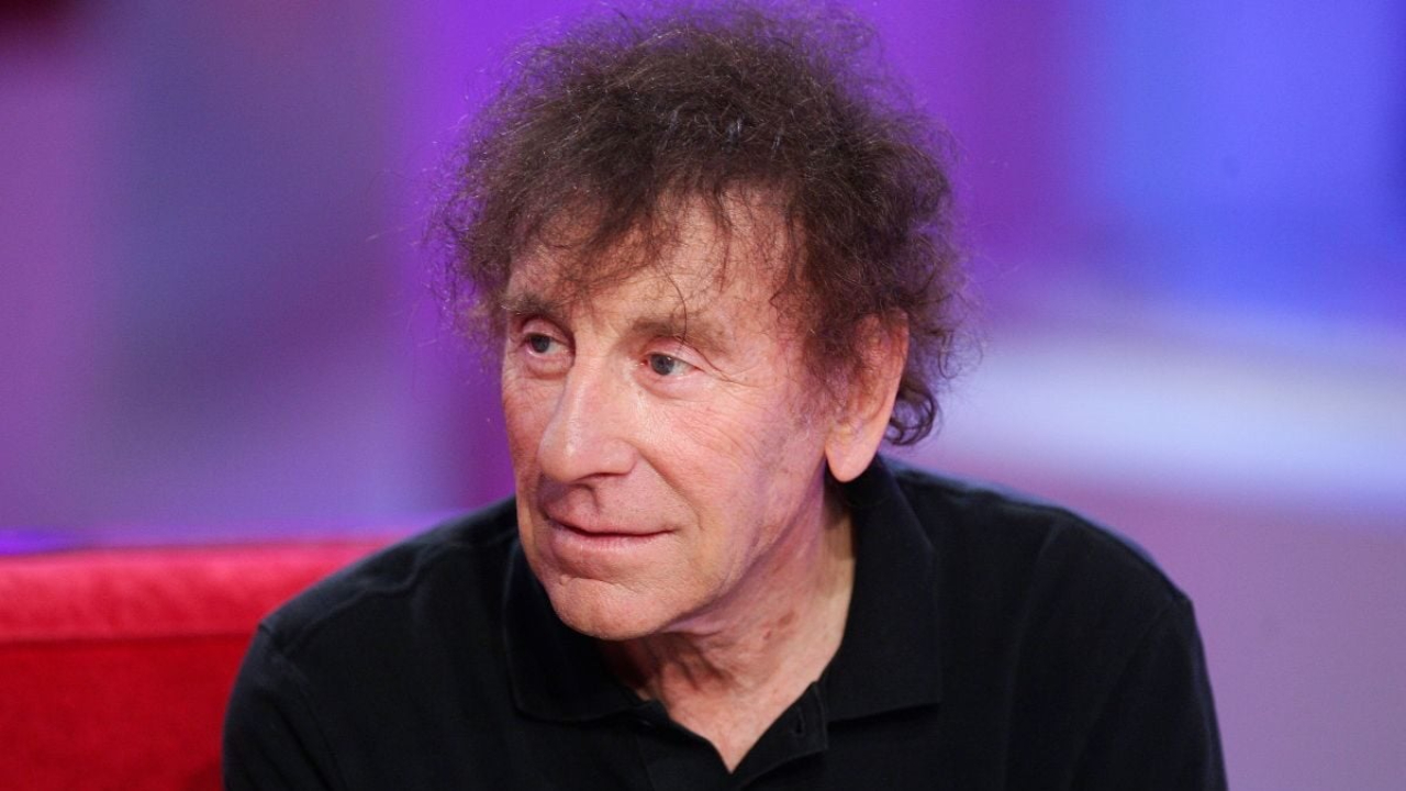 Portrait d’un homme aux cheveux bouclés grisonnants, assis sur un canapé rouge, éclairé par une lumière violette en studio, regard tourné vers la gauche.