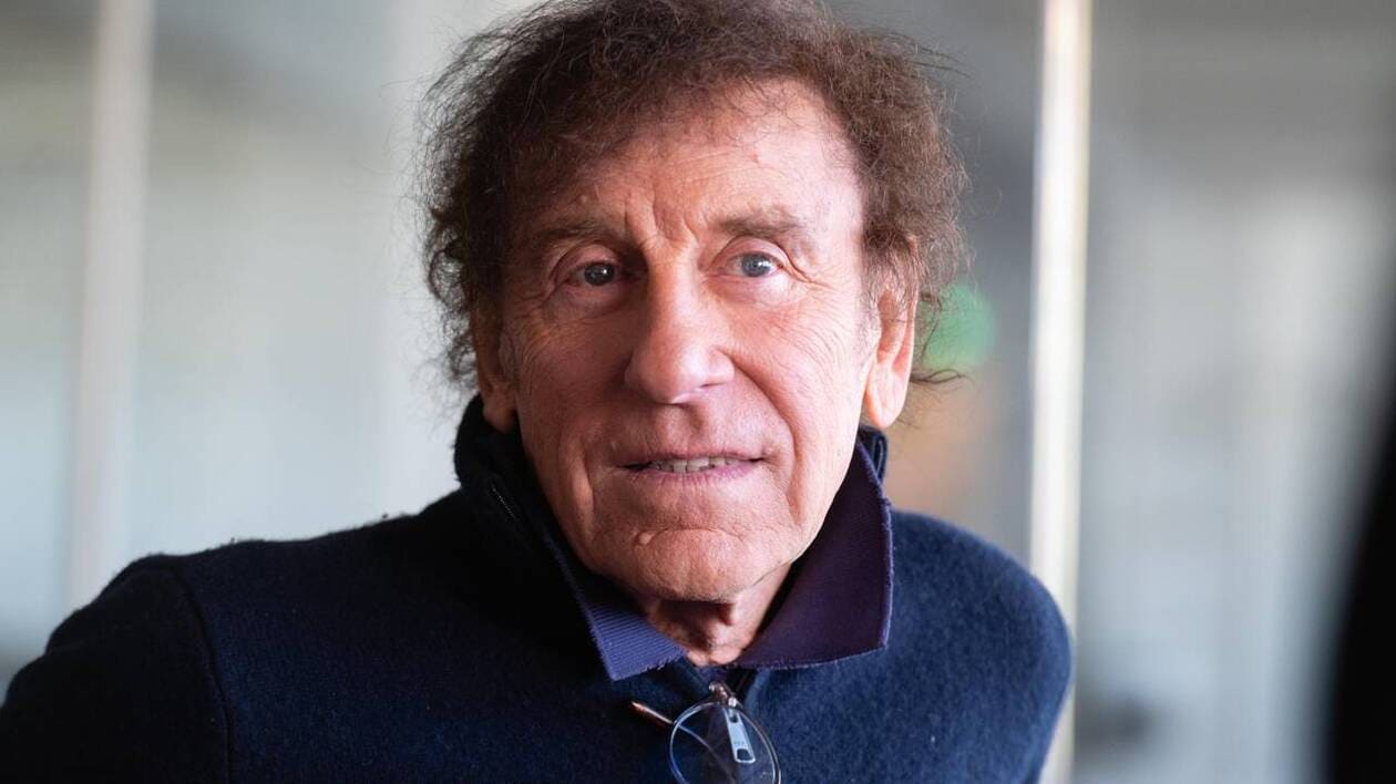 Portrait d’un homme aux cheveux bruns et bouclés, le visage marqué par l’âge, portant un manteau sombre, photographié en lumière naturelle avec un arrière-plan flou.