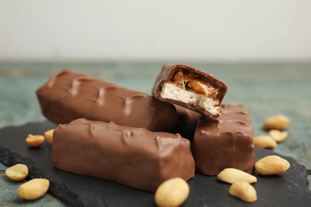 Barres chocolatées coupées montrant du caramel fondant et du nougat, posées sur une ardoise avec des cacahuètes autour.