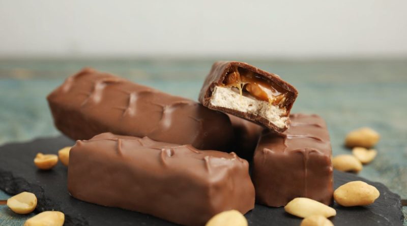 Barres chocolatées coupées montrant du caramel fondant et du nougat, posées sur une ardoise avec des cacahuètes autour.