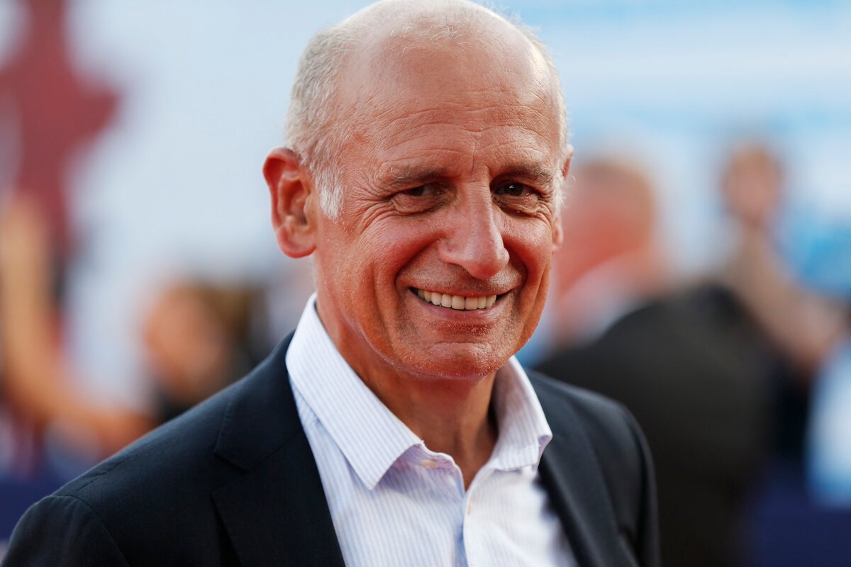 Homme chauve souriant en costume sombre et chemise claire, photographié en extérieur avec un fond flou de personnes et de lumière naturelle.