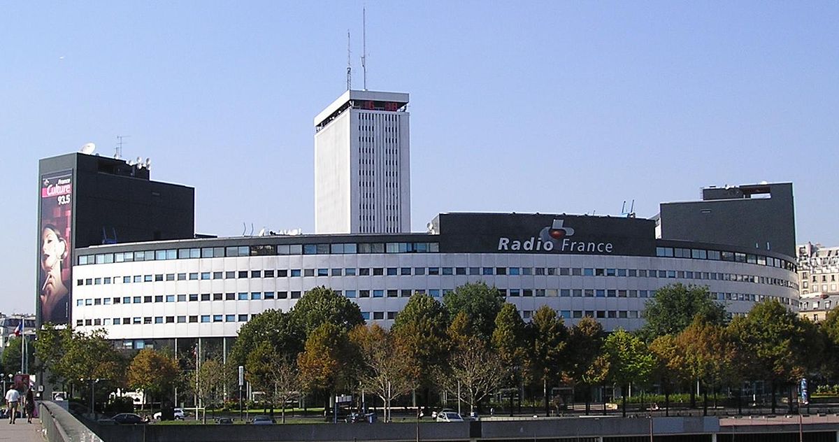 Façade de la Maison de la Radio à Paris, vaste bâtiment circulaire entouré d’arbres, avec ses tours et logos visibles sous un ciel clair.