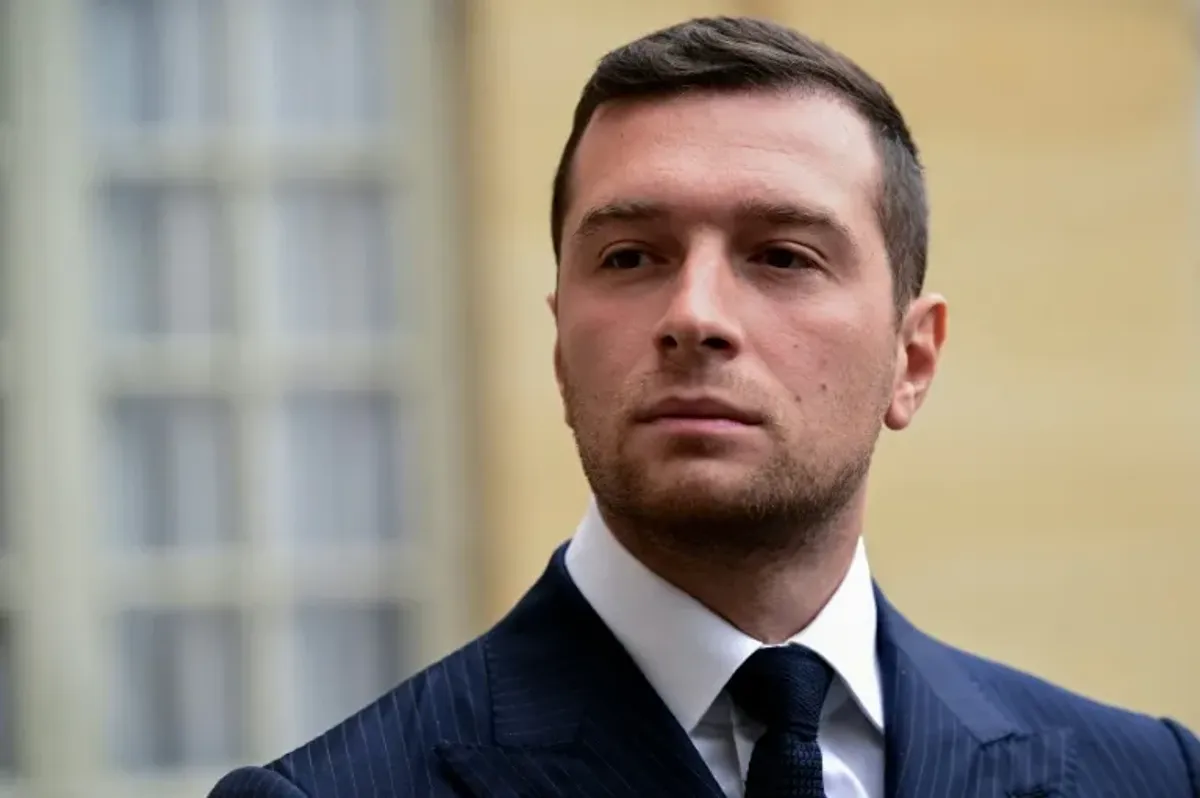 Homme en costume sombre et cravate, au visage sérieux, regardant légèrement sur le côté, en extérieur devant une façade aux grandes fenêtres.