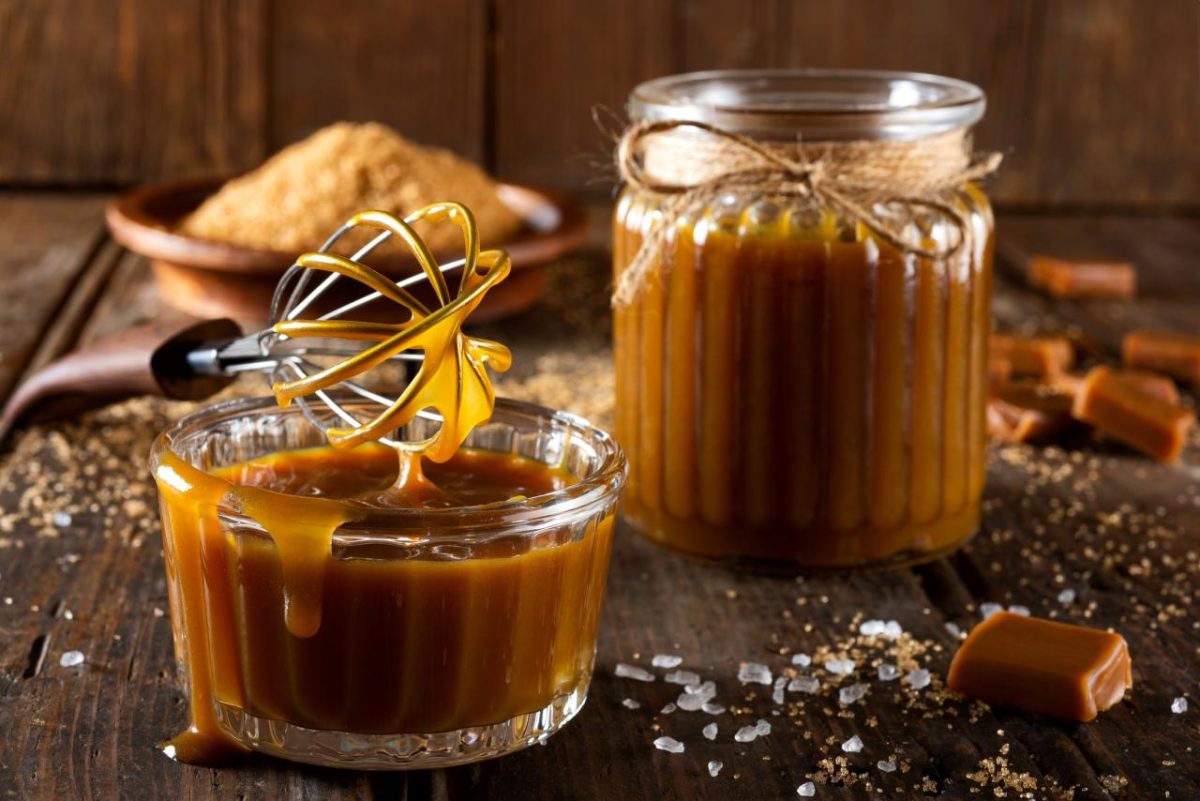 Caramel onctueux dans un petit bol en verre, nappé et brillant, avec un fouet dégoulinant de caramel, posé sur une table en bois rustique