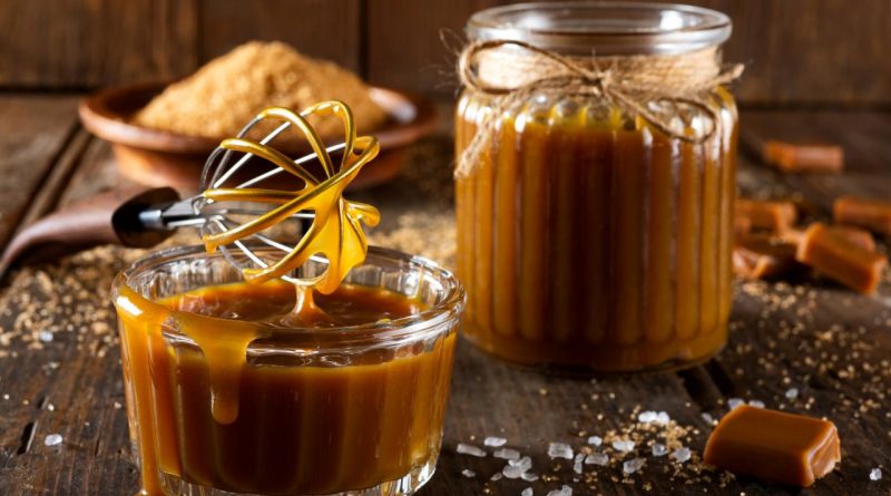 Caramel onctueux dans un petit bol en verre, nappé et brillant, avec un fouet dégoulinant de caramel, posé sur une table en bois rustique