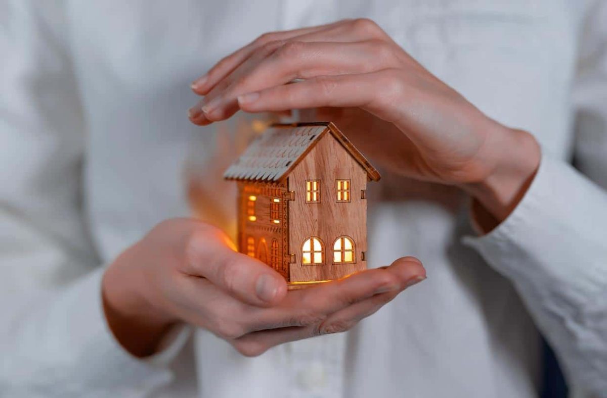 Personne tenant délicatement une petite maison en bois illuminée entre ses mains, symbole d’aide au logement et de soutien énergétique.