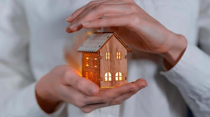 Personne tenant délicatement une petite maison en bois illuminée entre ses mains, symbole d’aide au logement et de soutien énergétique.