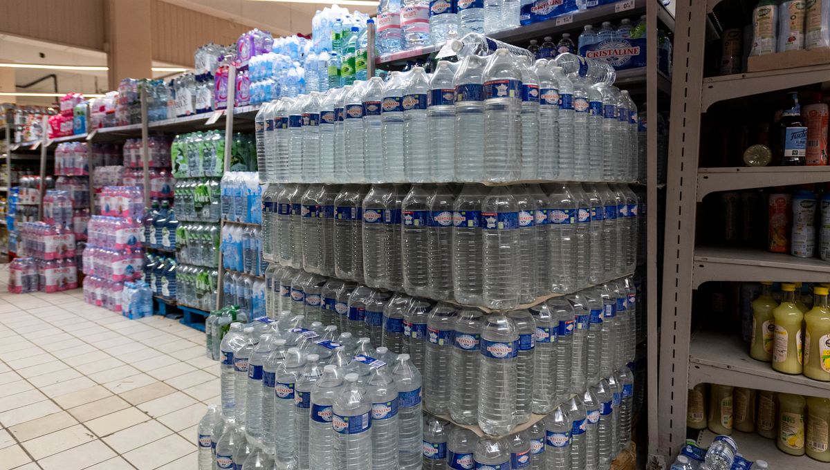 Rayons de supermarché remplis de bouteilles d’eau en plastique empilées sur des palettes et des étagères, illustrant la forte consommation et les enjeux autour du prix et du recyclage des emballages.