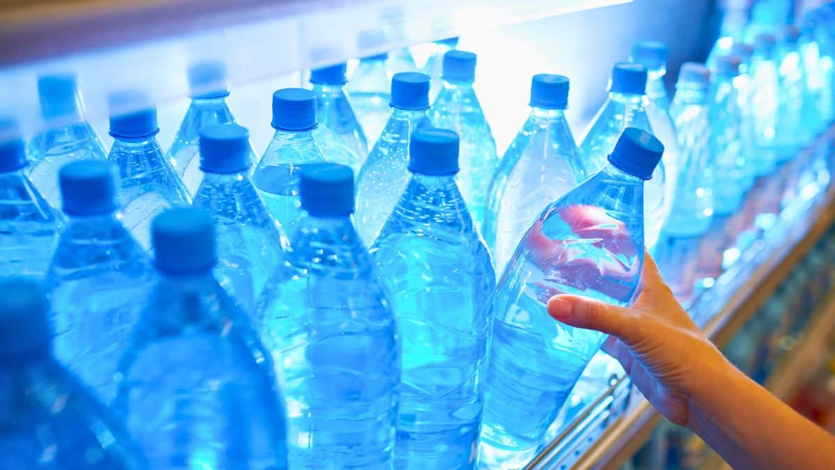 Main prenant une bouteille d’eau minérale parmi des dizaines d’autres alignées sur un rayon de supermarché, illustrant un rappel de produits et une alerte sanitaire concernant plusieurs marques d’eau en bouteille.