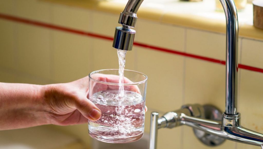Main tenant un verre transparent sous un robinet de cuisine en train de couler, illustrant la consommation d’eau du robinet et la question de sa qualité ou de sa pollution.