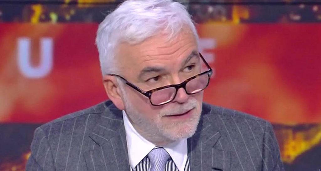 Homme aux cheveux blancs portant des lunettes et un costume rayé, assis sur un plateau de télévision avec un fond lumineux flou, en pleine discussion lors d’une émission d’actualité.