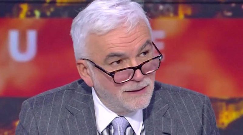Homme aux cheveux blancs portant des lunettes et un costume rayé, assis sur un plateau de télévision avec un fond lumineux flou, en pleine discussion lors d’une émission d’actualité.