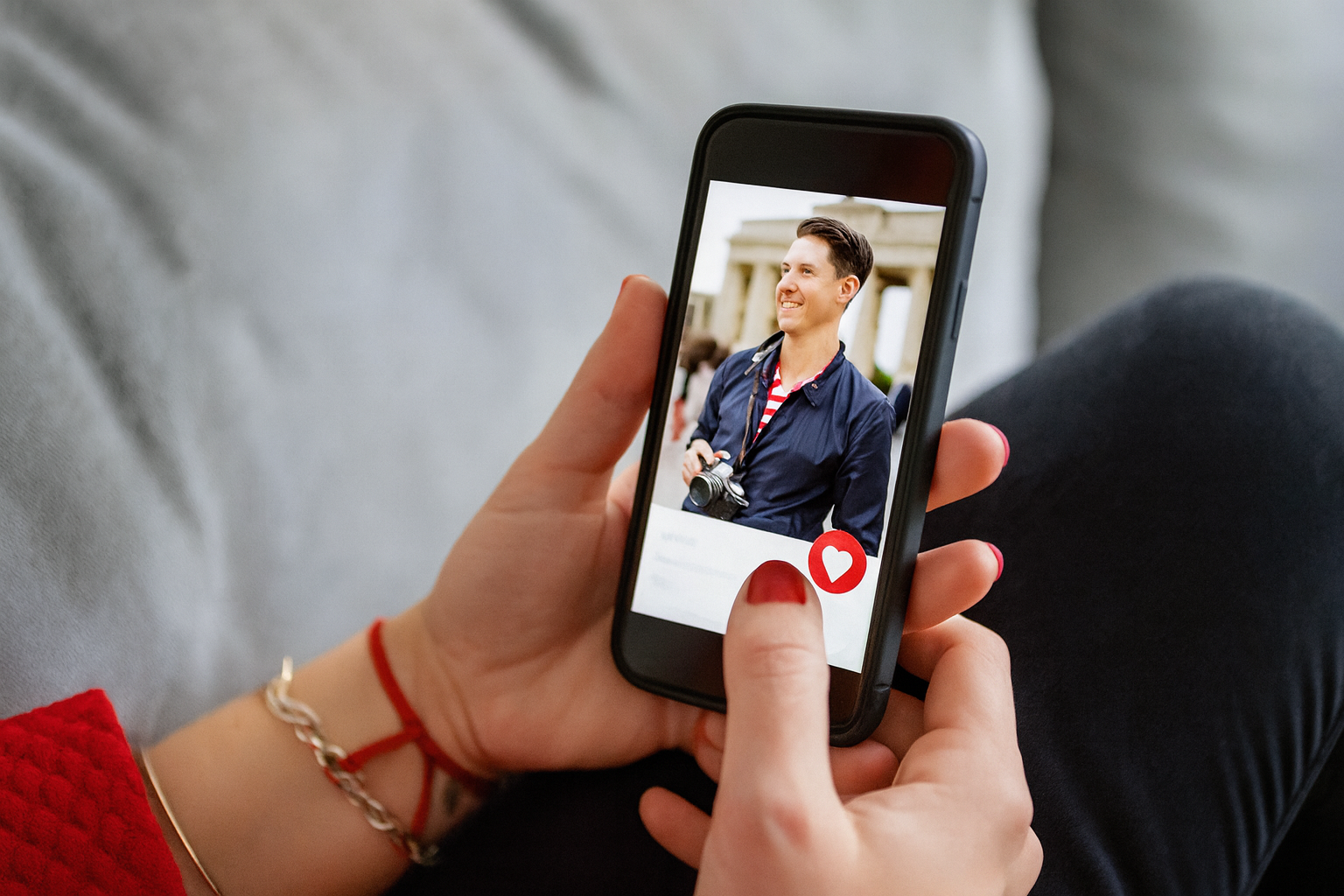 Main tenant un smartphone affichant un profil sur une application de rencontres, avec un cœur en bas de l’écran, en situation de navigation sur canapé.
