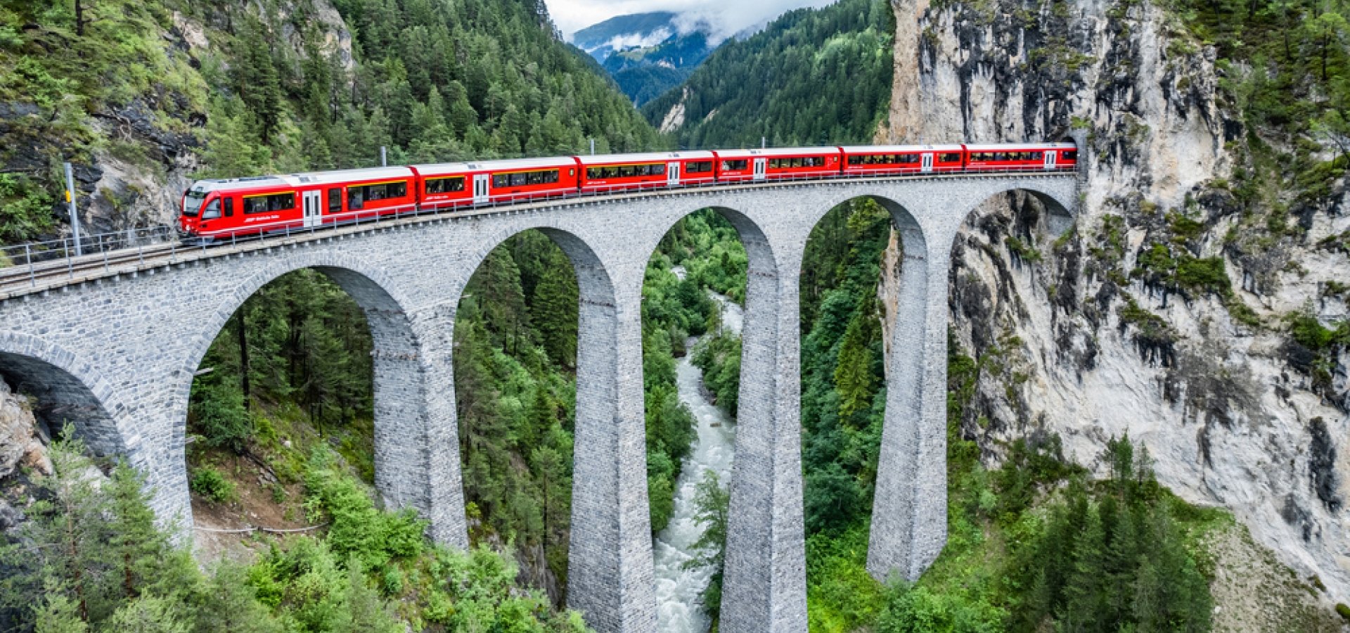 Train rouge traversant un haut viaduc en pierre au-dessus d’une vallée boisée, entouré de montagnes rocheuses et de forêts denses.