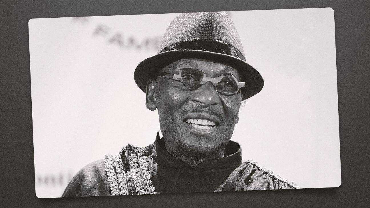 Portrait de Jimmy Cliff souriant, debout sur scène, baigné de lumière, avec un arrière-plan flou rappelant l’ambiance d’un concert reggae.