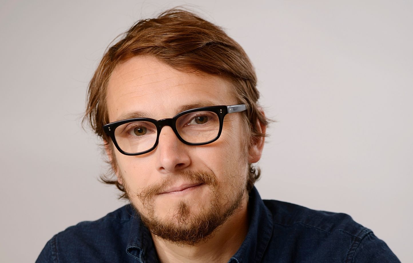 Portrait d’un homme brun portant des lunettes noires épaisses, barbe légère, expression neutre, photographié en gros plan sur un fond clair.