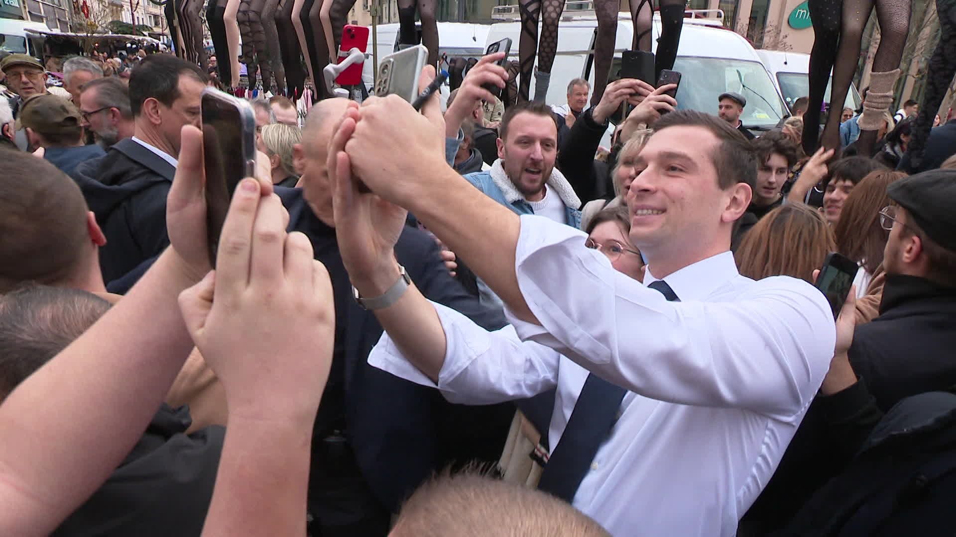 Scène de foule dans une foire : un homme en chemise blanche sourit en prenant un selfie entouré de nombreux visiteurs levant leurs téléphones, ambiance populaire et animée en extérieur.
