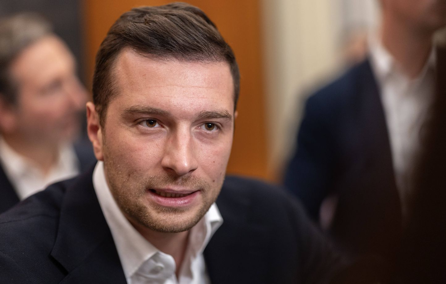 Portrait d’un homme en veste sombre et chemise blanche, filmé en gros plan dans un environnement intérieur, au milieu d’autres personnes légèrement floutées à l’arrière-plan.