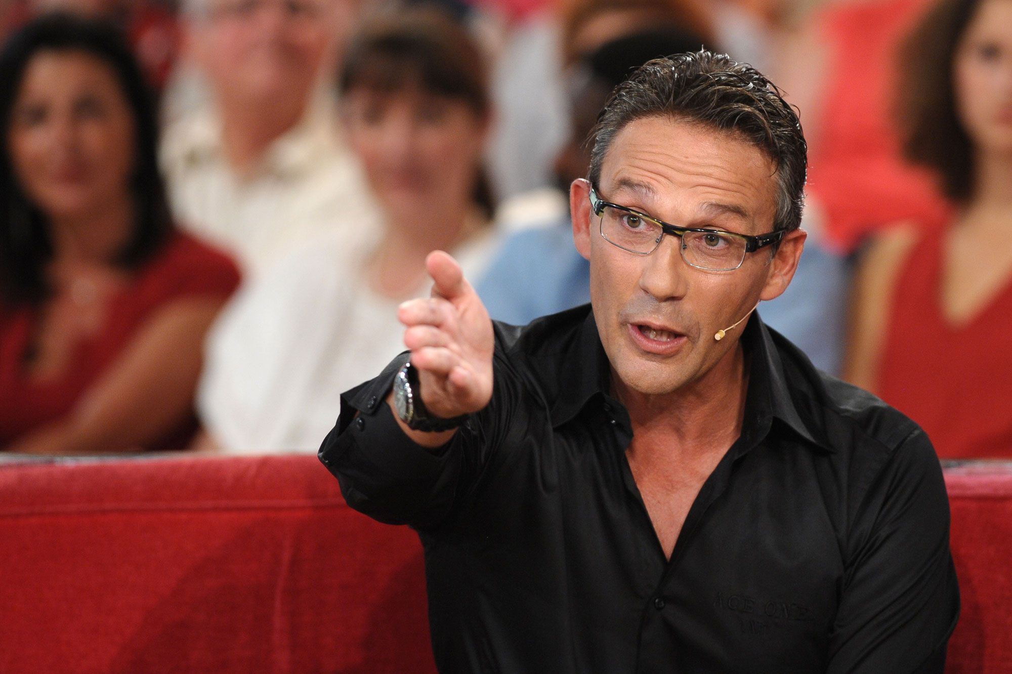 Animateur de télévision assis sur un plateau, vêtu d’une chemise noire, parlant avec énergie devant un public flou en arrière-plan.