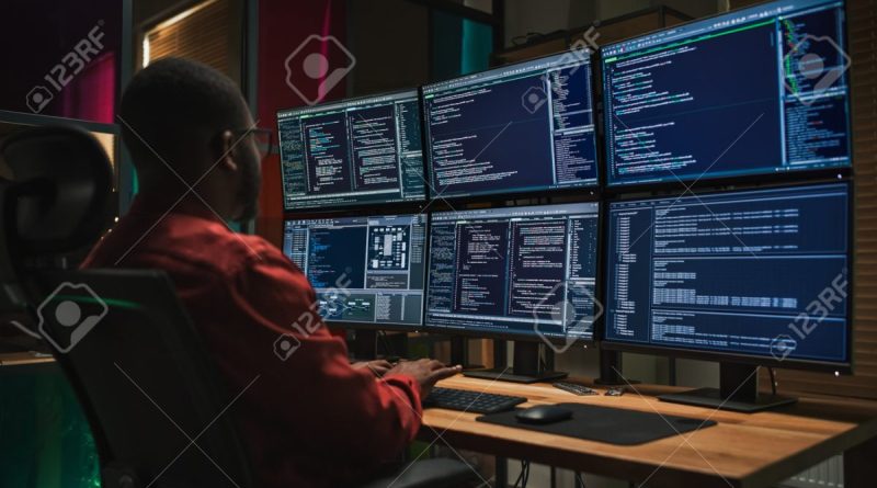 Homme assis devant plusieurs écrans d’ordinateur affichant des lignes de code, représentant une enquête informatique ou une activité de cybersécurité dans un bureau sombre.