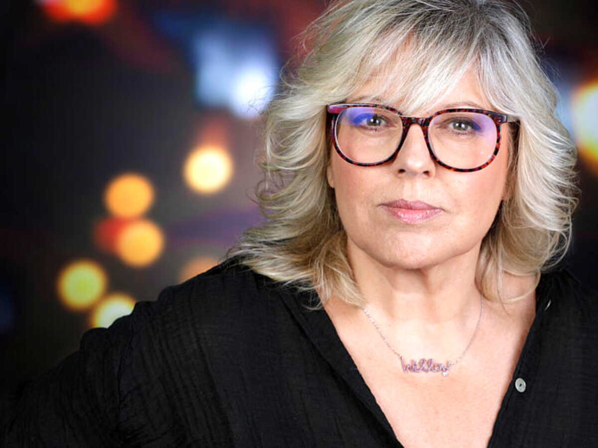 Femme aux cheveux blonds et lunettes à monture épaisse, regard déterminé face à l’objectif, sur fond flou de lumières colorées évoquant un plateau de télévision.