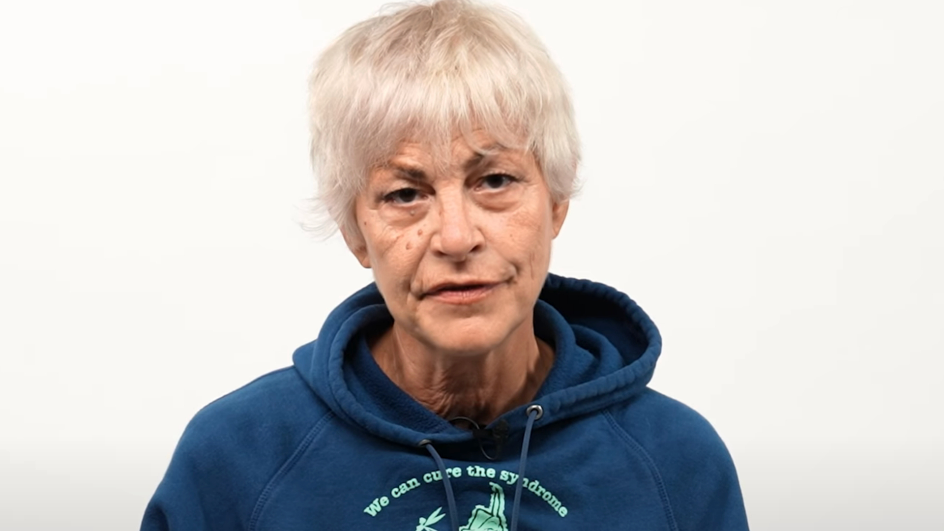 Femme aux cheveux courts gris, portant un sweat à capuche bleu, filmée en gros plan devant un fond blanc, avec une expression sérieuse.