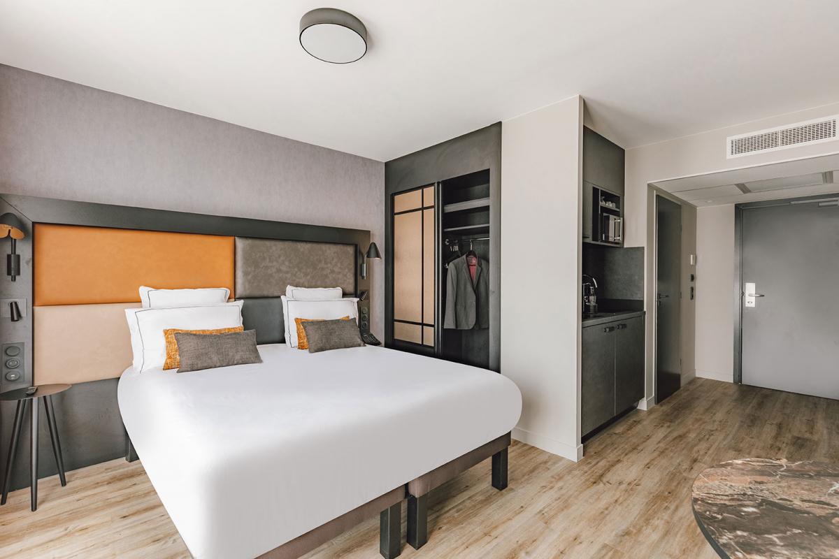 Chambre moderne et lumineuse avec un grand lit double aux coussins contrastés, un dressing ouvert contenant quelques vêtements, un coin kitchenette sombre et un sol en parquet clair.