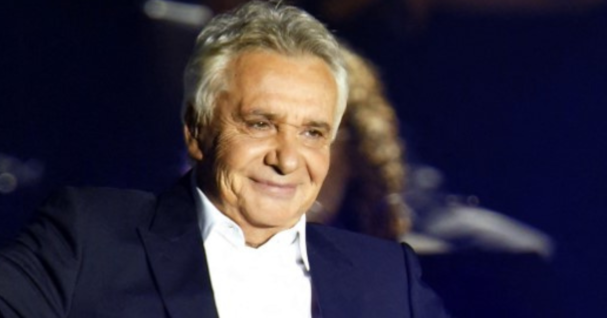 Un homme aux cheveux gris, souriant sur scène, vêtu d’un costume sombre sous un éclairage de concert.