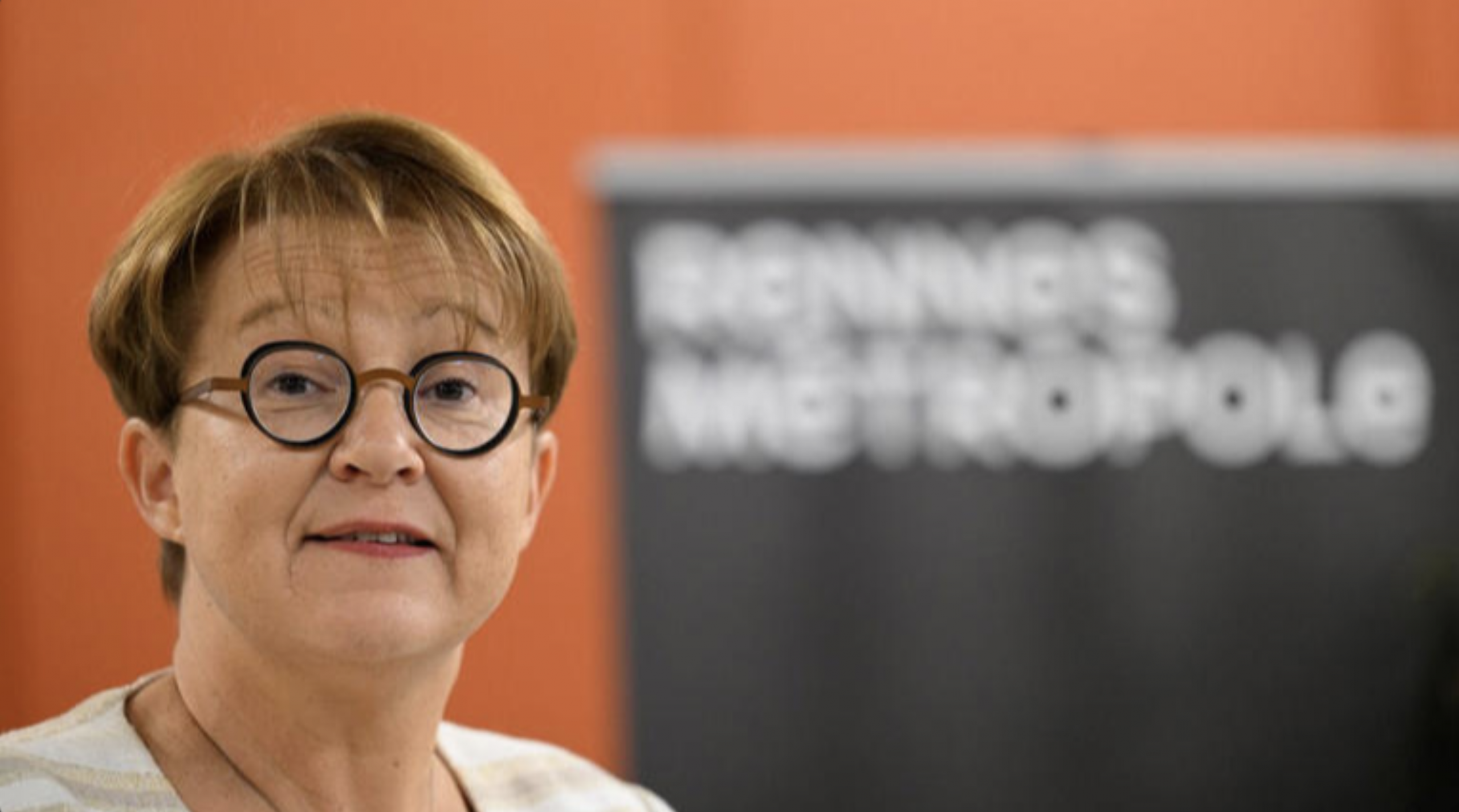 Portrait d’une femme portant des lunettes rondes, debout devant un fond orange avec un panneau flou indiquant « Rennes Métropole ».