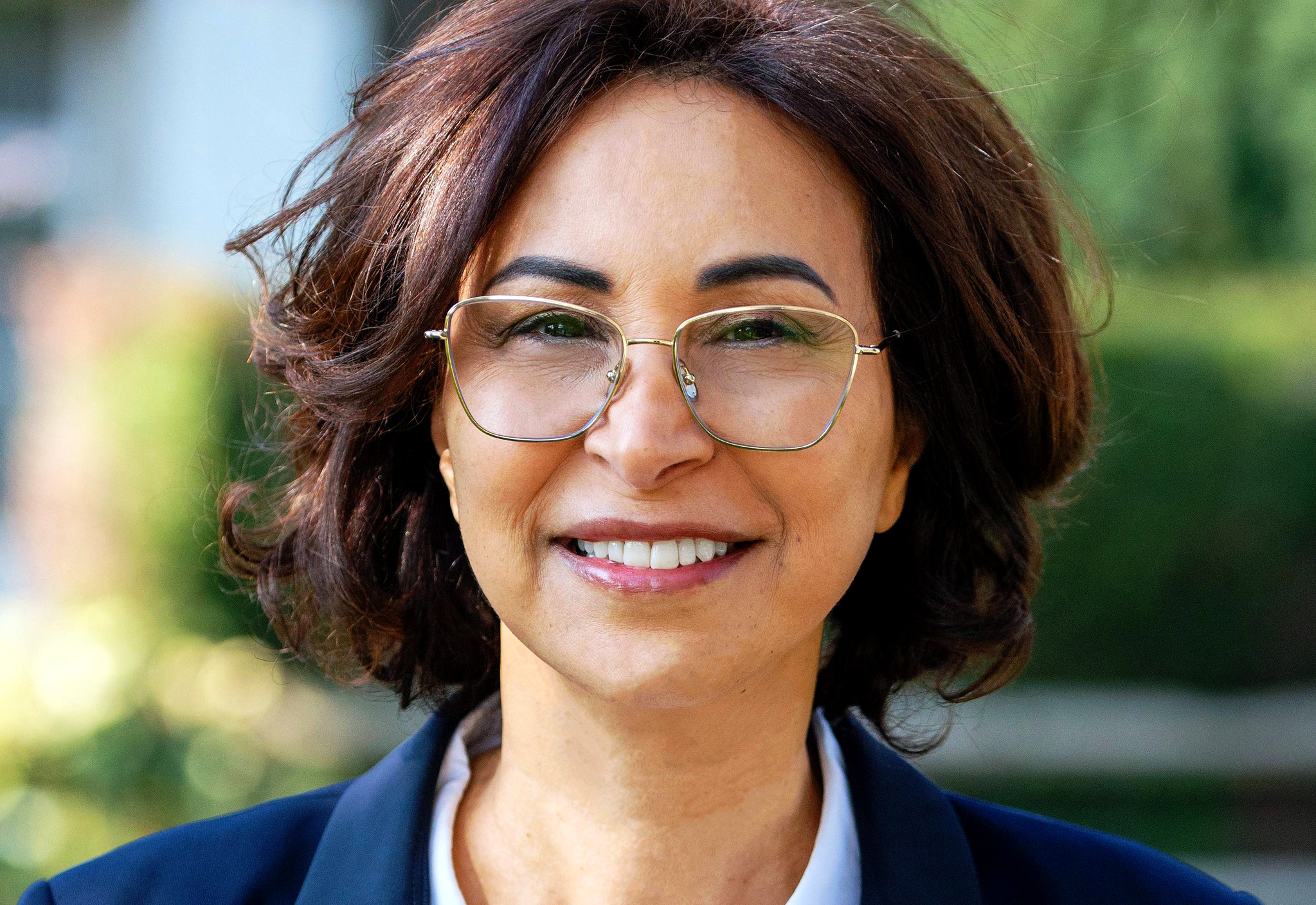 Portrait en gros plan d’une femme souriante portant des lunettes à monture dorée, les cheveux bruns légèrement ondulés, photographiée en extérieur avec un arrière-plan flou de verdure.