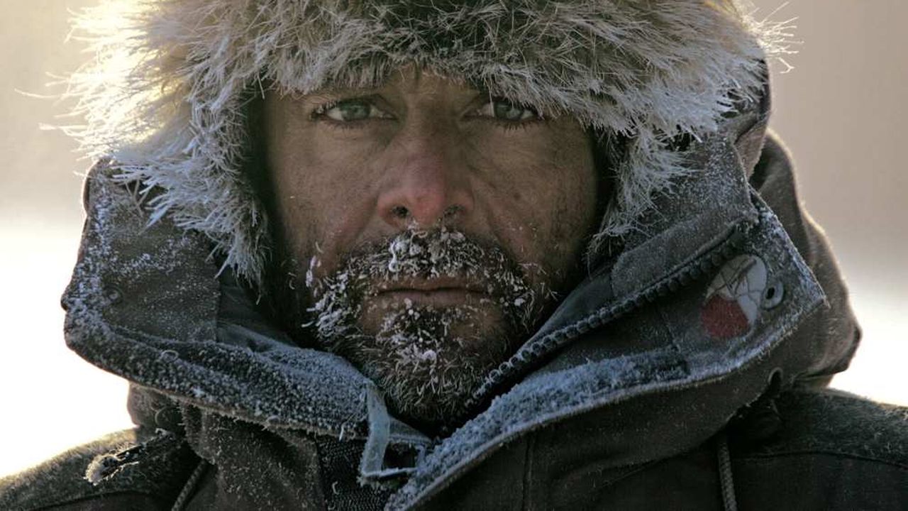 Portrait rapproché d’un explorateur en parka polaire, le visage couvert de givre et de neige, regard intense sous une capuche bordée de fourrure dans un environnement glacé.