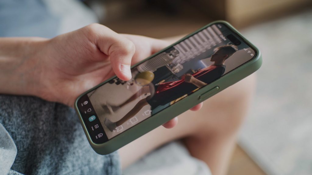 Main tenant un smartphone affichant une vidéo en plein écran, montrant un jeune homme dans une pièce éclairée, scène vue de près et en légère contre-plongée.