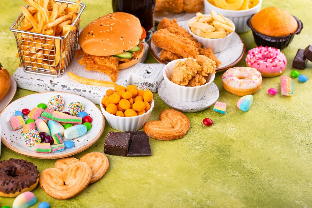 Assortiment d’aliments ultra-transformés et très sucrés, comprenant burgers, frites, poulet frit, donuts, biscuits et bonbons, disposés sur une table pour illustrer les pièges alimentaires soi-disant sains.