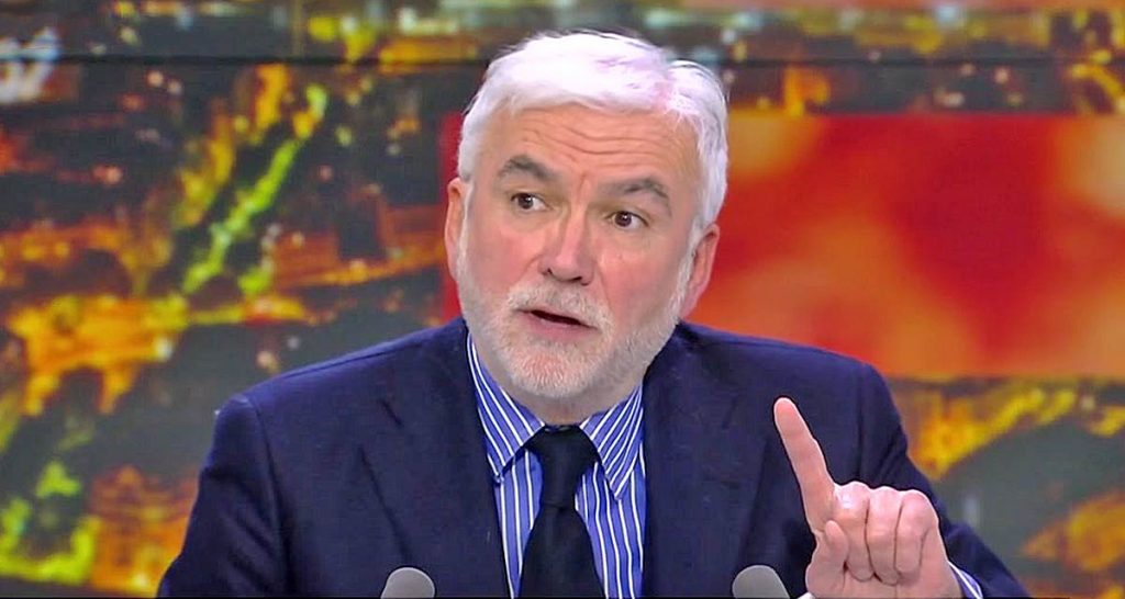 Homme aux cheveux gris et à la barbe blanche, en costume sombre et chemise à rayures, parlant en plateau TV et levant légèrement l’index devant un décor lumineux flou.